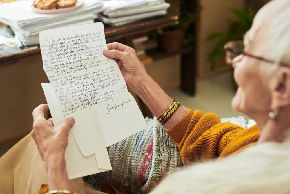 Elderly person reading an intricately handwritten letter, sitting beside a cozy desk filled with stacked papers and plants, expressing emotions of nostalgia and longing while reminiscing