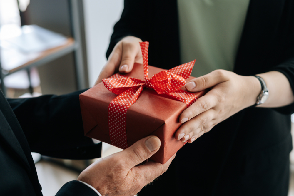 Close-up view of hands of unrecognizable woman giving red gift box tied to bow handed to man. Giving gifts during the Christmas, Happy New Year and Happy Birthday at office